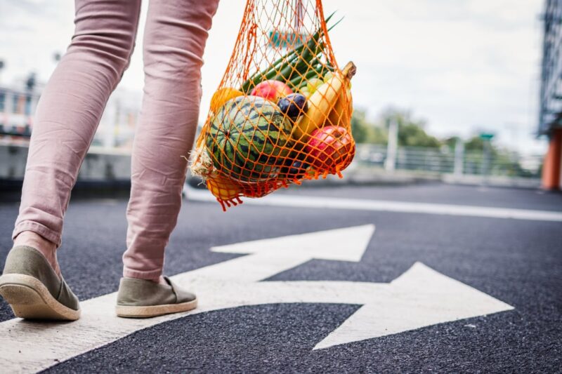 A person walking while carrying a reusable mesh bag filled with fresh fruits and vegetables, symbolizing healthy, sustainable food choices.