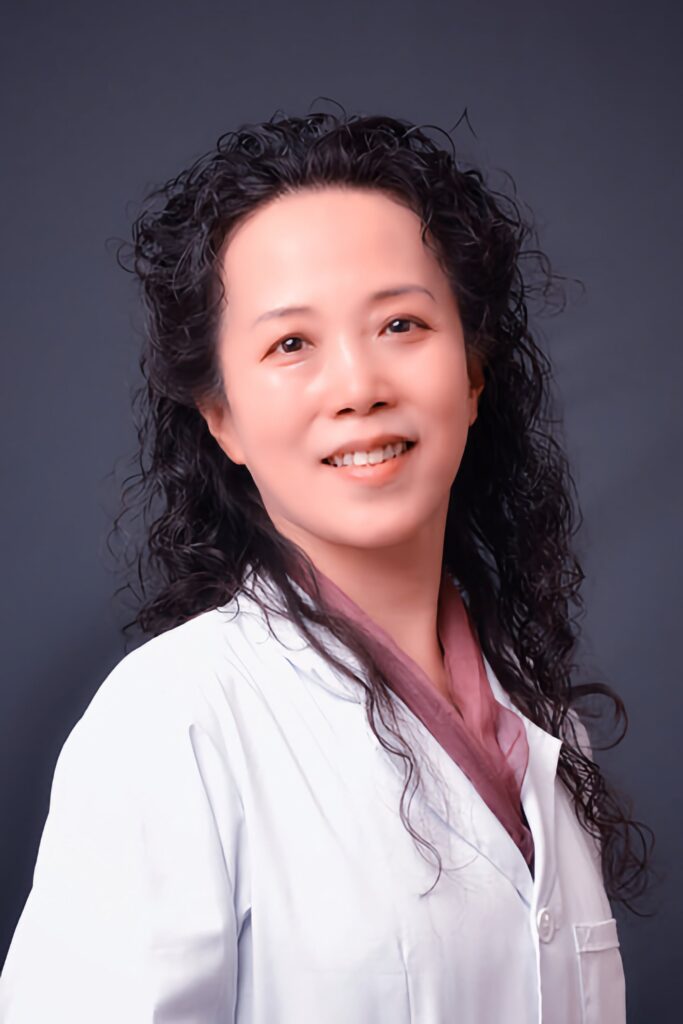 A smiling female practitioner in a white lab coat, with long, curly hair, standing against a dark background, exuding a professional and approachable demeanor.