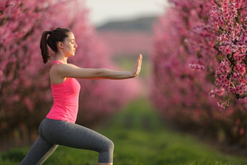 A woman practicing Qi Gong in a peaceful outdoor setting, surrounded by blooming pink trees, focusing on gentle movements and breath control to cultivate inner balance and energy flow.