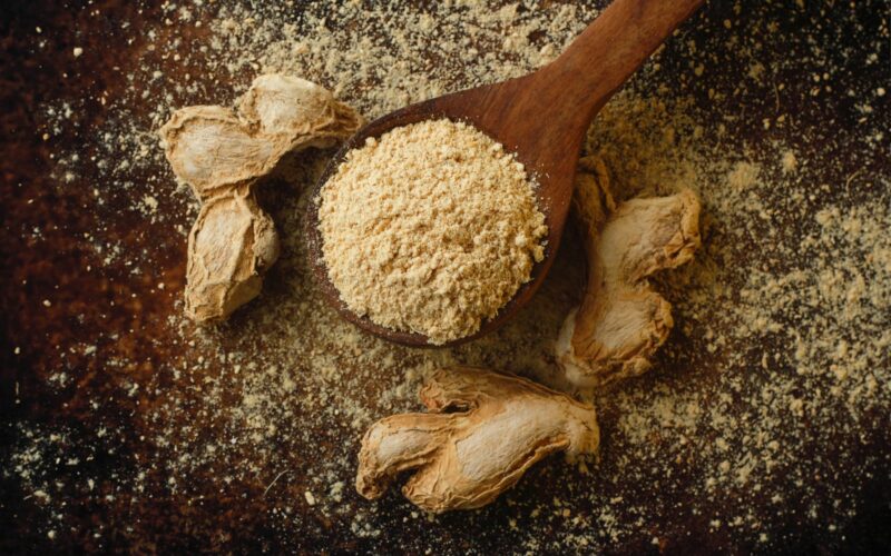 wooden spoon filled with ground ginger surrounded by dried ginger roots, symbolizing traditional herbal remedies